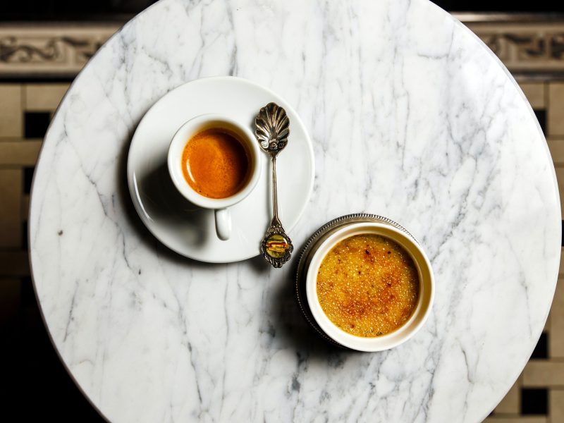 Creme brulee dessert and a cup of espresso with a beautiful old spoon on a marble table, top view